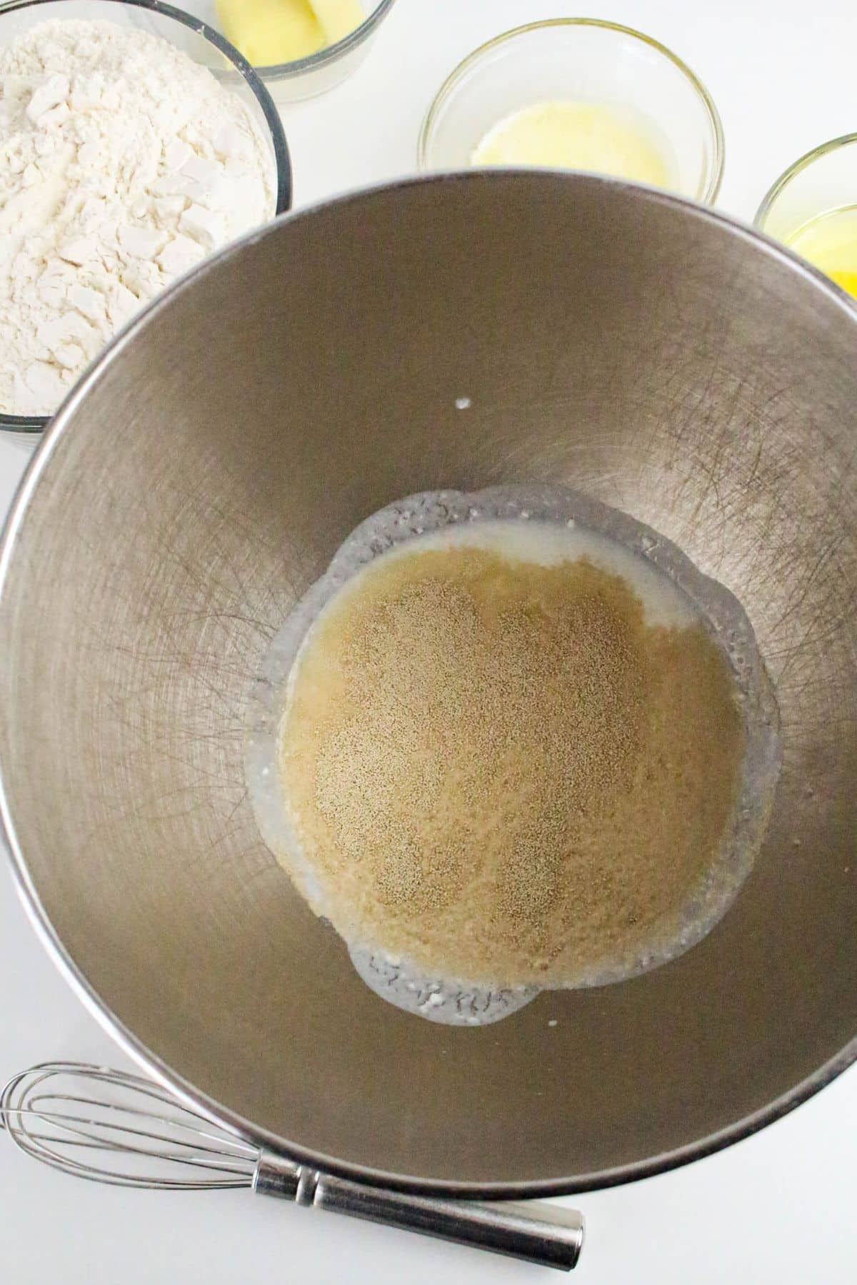 A mixing bowl containing yeast and liquid, surrounded by bowls of flour, melted butter, eggs, and a whisk on a white surface.