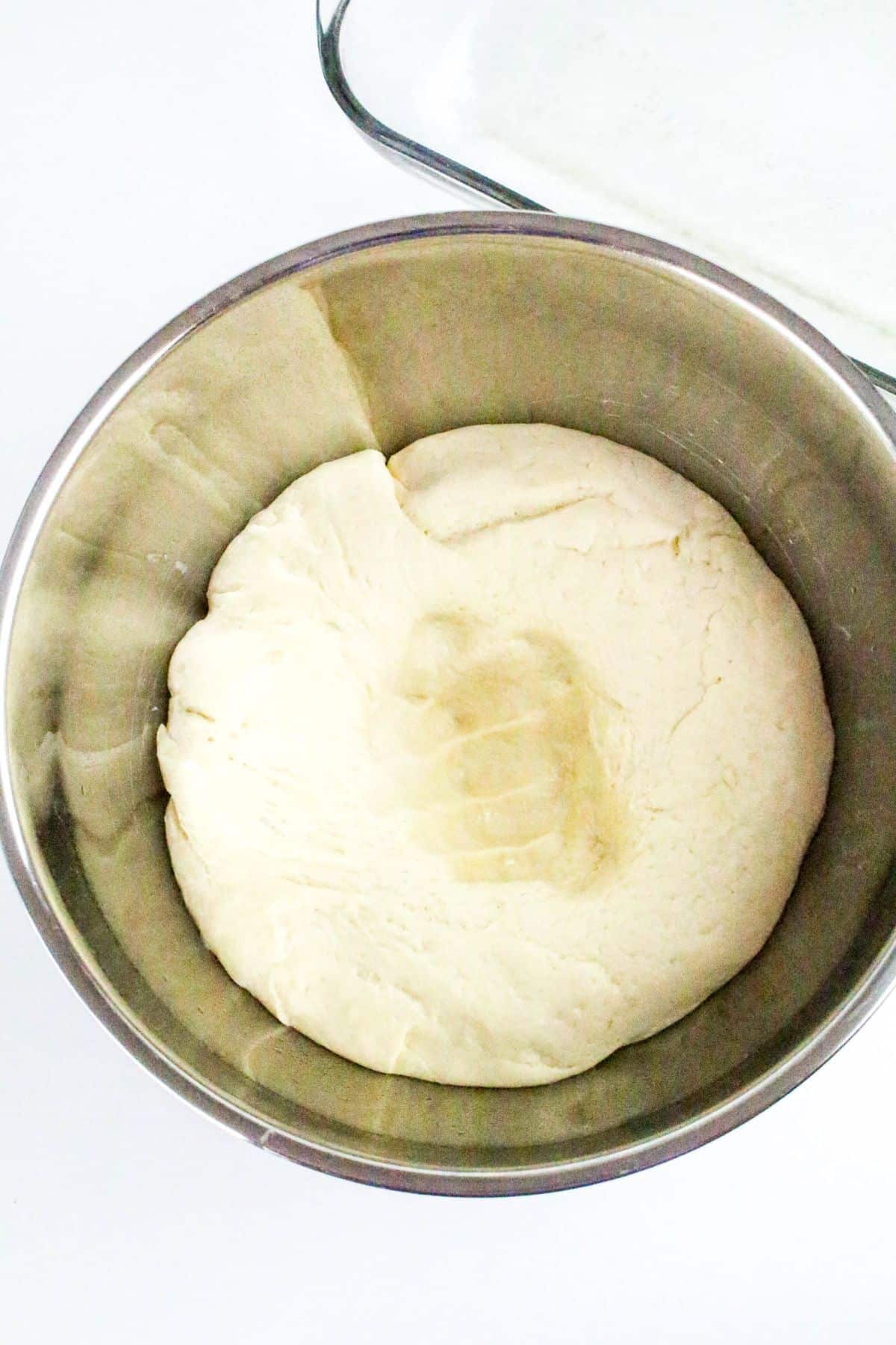 A stainless steel bowl containing risen dough with a handprint on top, placed on a white surface.