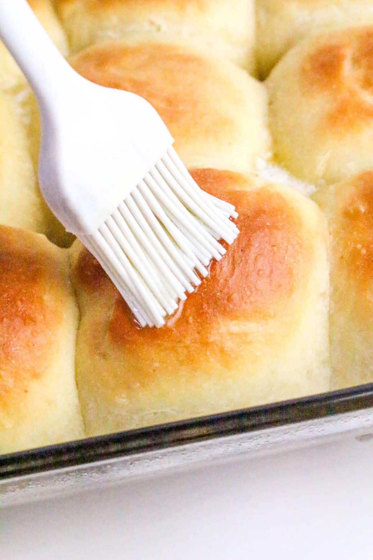 A pastry brush is being used to coat freshly baked golden brown dinner rolls in a baking dish.