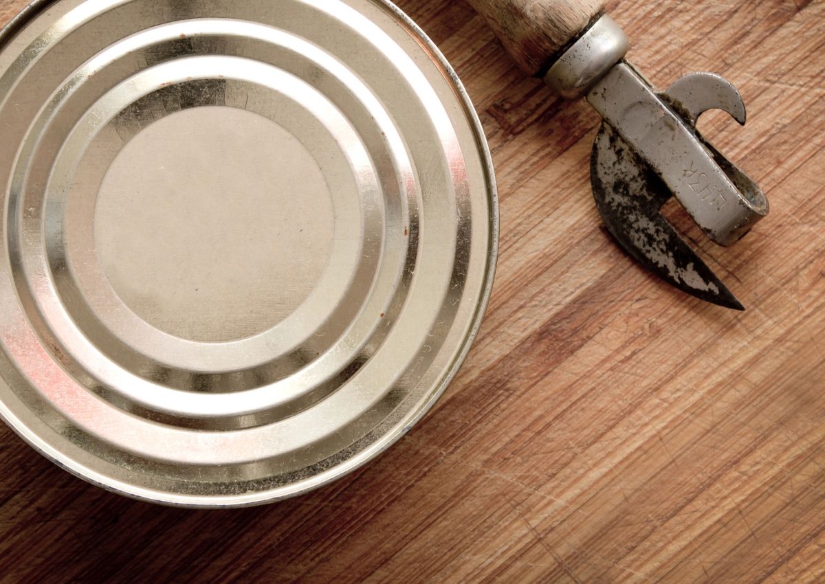 A closed tin can and a vintage manual can opener rest on a wooden surface.