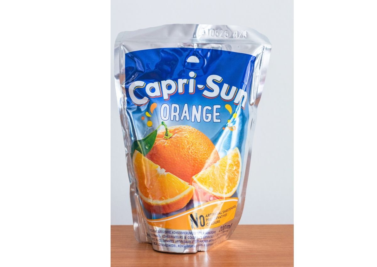 A Capri-Sun Orange juice pouch stands on a wooden surface, displaying orange fruit images and product branding on the front.
