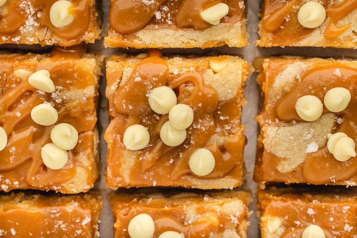 Close-up of caramel blondie bars topped with caramel sauce, white chocolate chips, and a sprinkle of sea salt, arranged in a grid pattern.