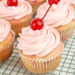 Pink frosted cupcakes topped with cherries are arranged on a metal cooling rack.