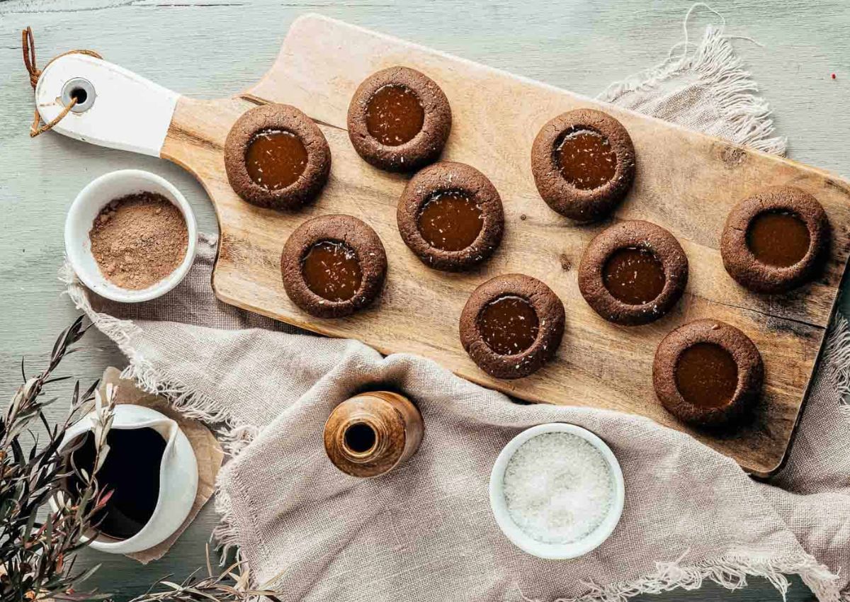 A wooden board with nine chocolate thumbprint cookies filled with caramel, next to bowls of cocoa powder, coarse salt, and a small jar of syrup on a cloth.