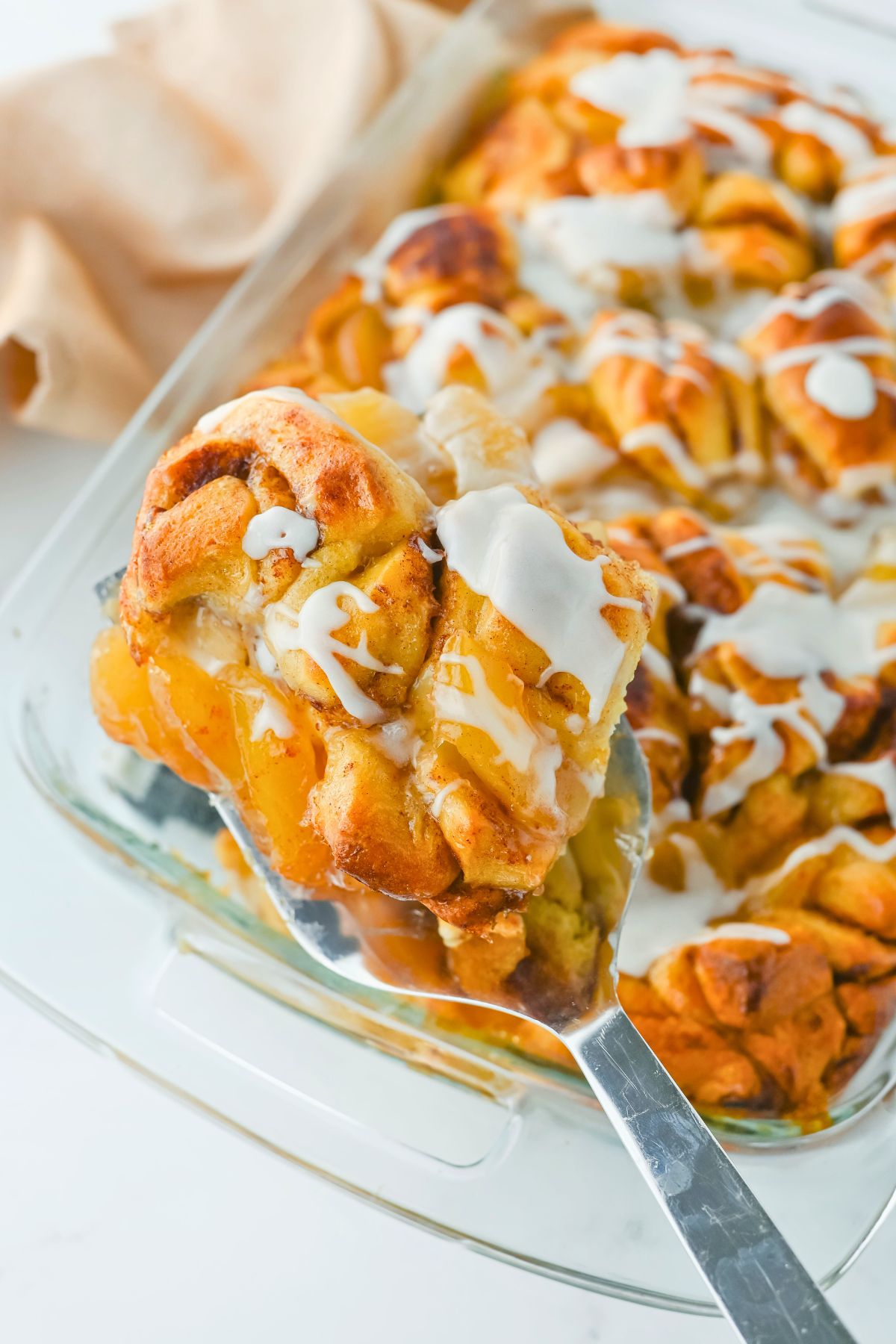 A metal spatula lifts a frosted cinnamon roll from a glass baking dish filled with cinnamon rolls topped with white icing.
