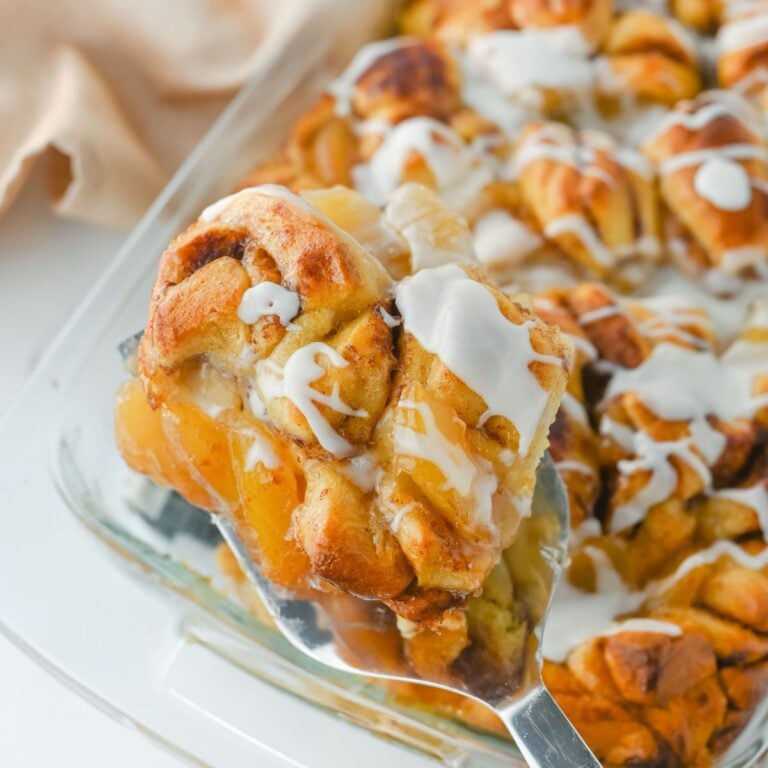 A close-up of a serving spatula lifting a glazed cinnamon roll from a glass baking dish filled with cinnamon rolls.