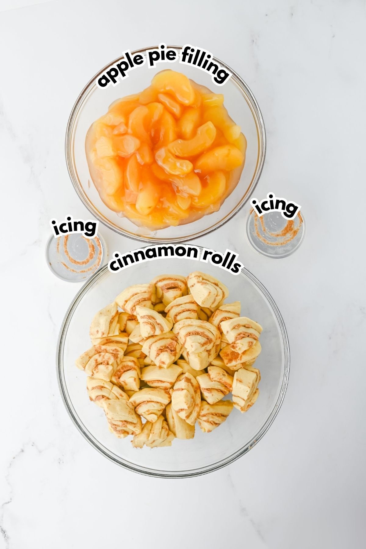 Two bowls on a white surface: one with apple pie filling, the other with cut cinnamon rolls, with two small containers of icing beside them. Labels identify each item.