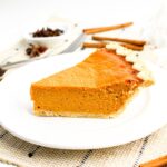 A slice of pumpkin pie on a white plate, with cinnamon sticks and star anise in the background.