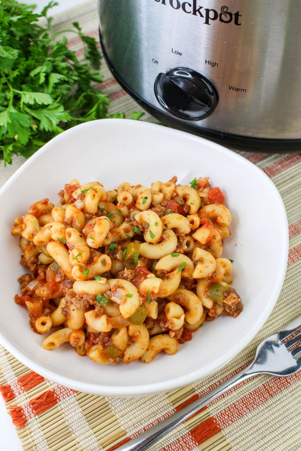 A white bowl of macaroni pasta with ground beef, tomatoes, and herbs sits on a placemat near a slow cooker and a bunch of fresh parsley.