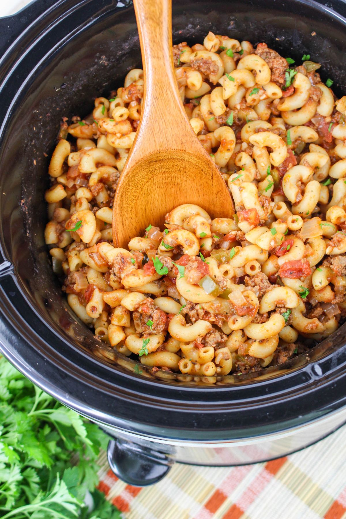 A wooden spoon stirs macaroni with ground meat and tomato sauce inside a slow cooker, with fresh parsley and a striped cloth nearby.