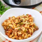A white bowl filled with macaroni, ground beef, diced tomatoes, and green peppers, garnished with parsley, sits on a placemat near a slow cooker.