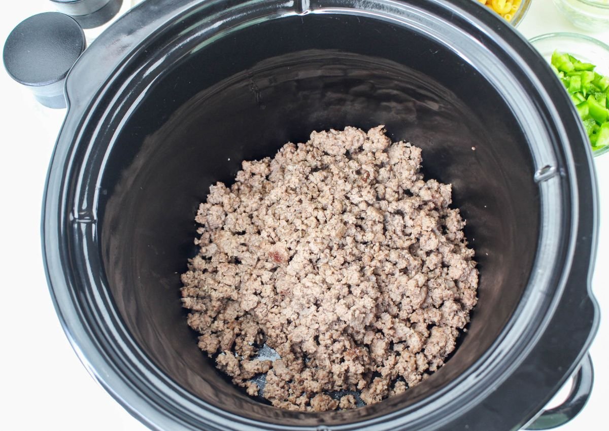 Cooked ground beef in the bottom of a black slow cooker, with bowls of chopped green peppers and other ingredients nearby.