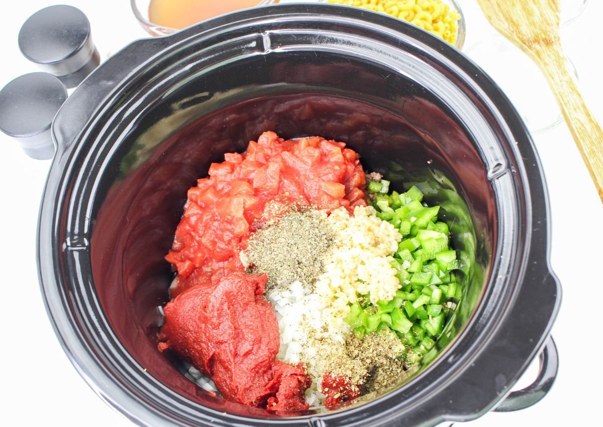 A slow cooker containing uncooked diced tomatoes, tomato paste, chopped green pepper, onions, garlic, and herbs, ready to be mixed. Macaroni and broth are visible in the background.