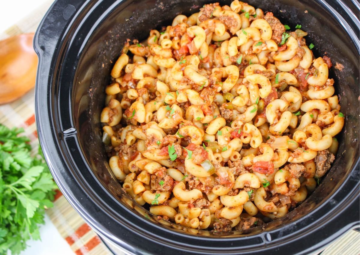 Cooked macaroni with ground beef, tomato sauce, and herbs in a black slow cooker, placed on a striped cloth with fresh parsley nearby.
