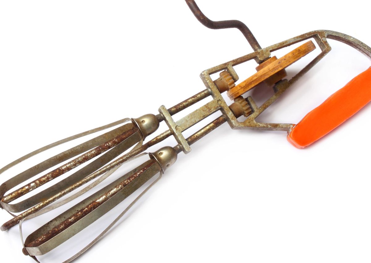Metal hand-crank egg beater with gears and an orange handle, showing signs of rust and wear, on a white background.