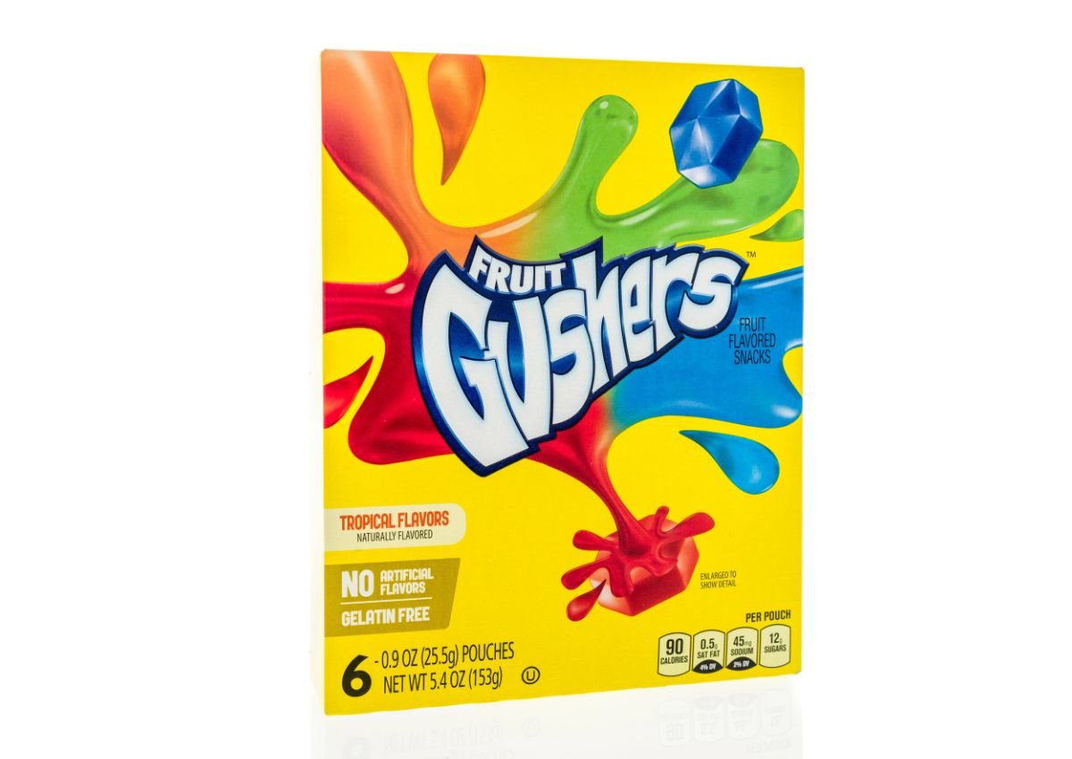 A box of Fruit Gushers fruit-flavored snacks in tropical flavors, featuring colorful splashes and a blue gem design on a yellow background.