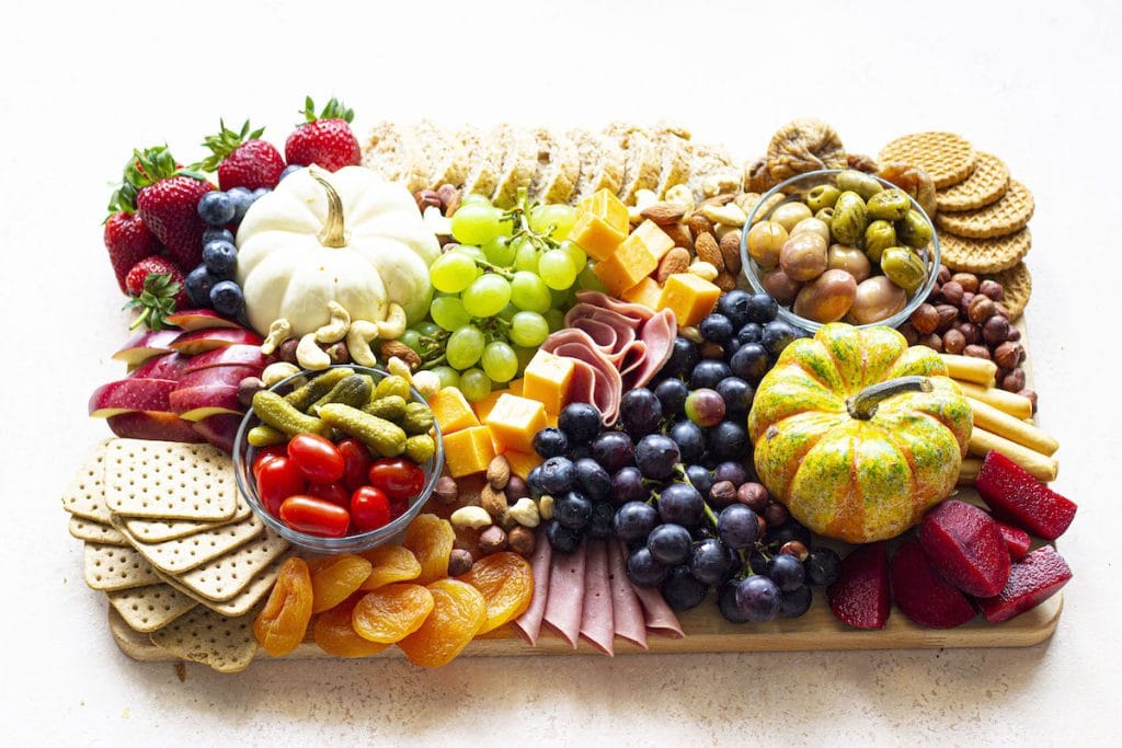 A charcuterie board with assorted cheeses, meats, crackers, fruits, nuts, olives, pickles, and two small decorative gourds arranged on a wooden tray.