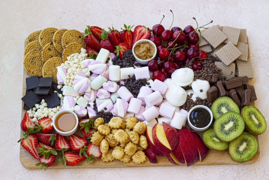 A wooden board topped with sliced strawberries, kiwi, apple, cherries, assorted cookies, mini crackers, marshmallows, chocolate pieces, dipping sauces, and wafer biscuits.