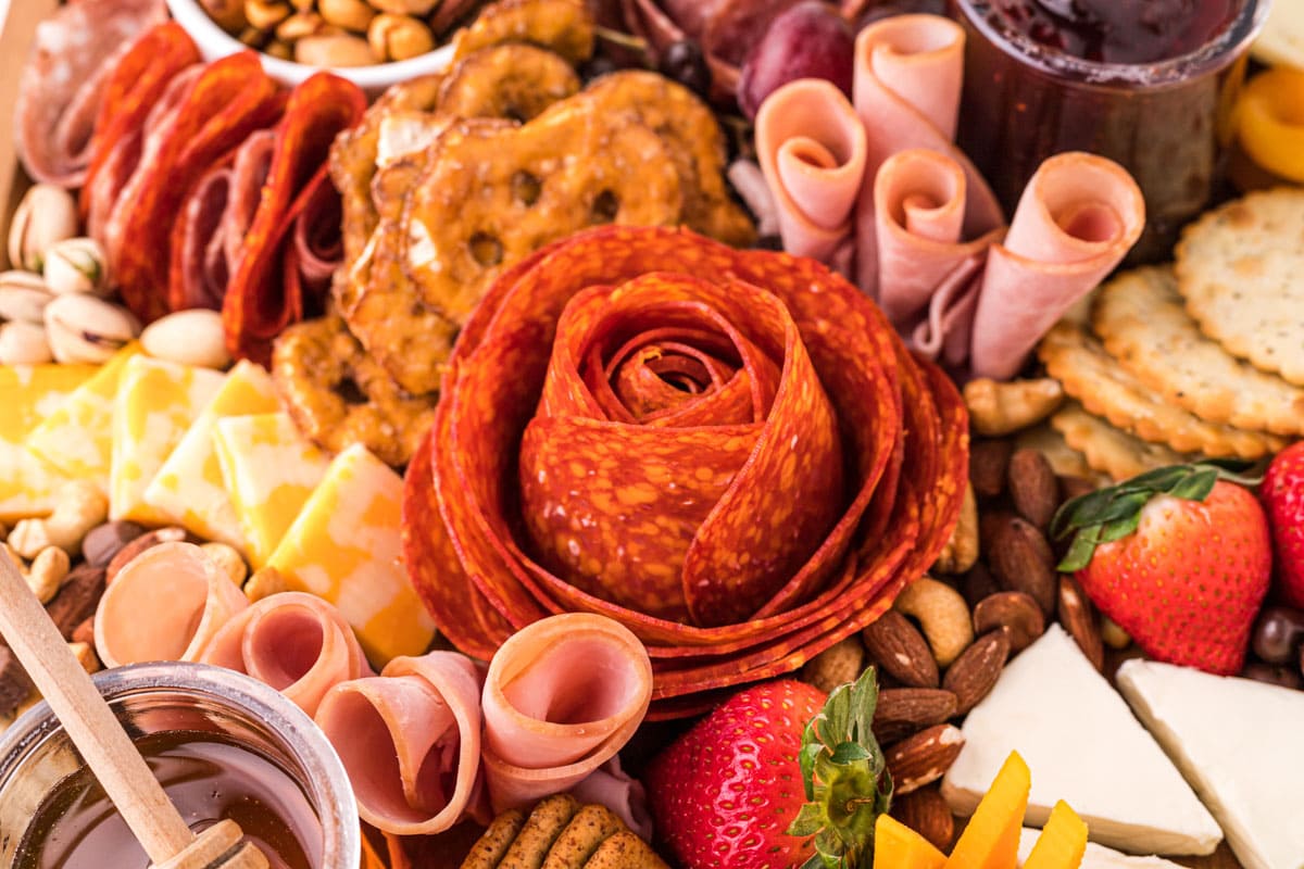 Charcuterie board with a salami rose centerpiece, assorted meats, cheeses, crackers, pretzels, nuts, strawberries, and jars of spreads.