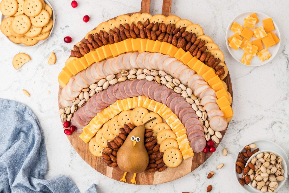 A round wooden board with rows of crackers, meats, cheeses, nuts, and a pear arranged to resemble a turkey, surrounded by small plates of snacks on a white countertop.