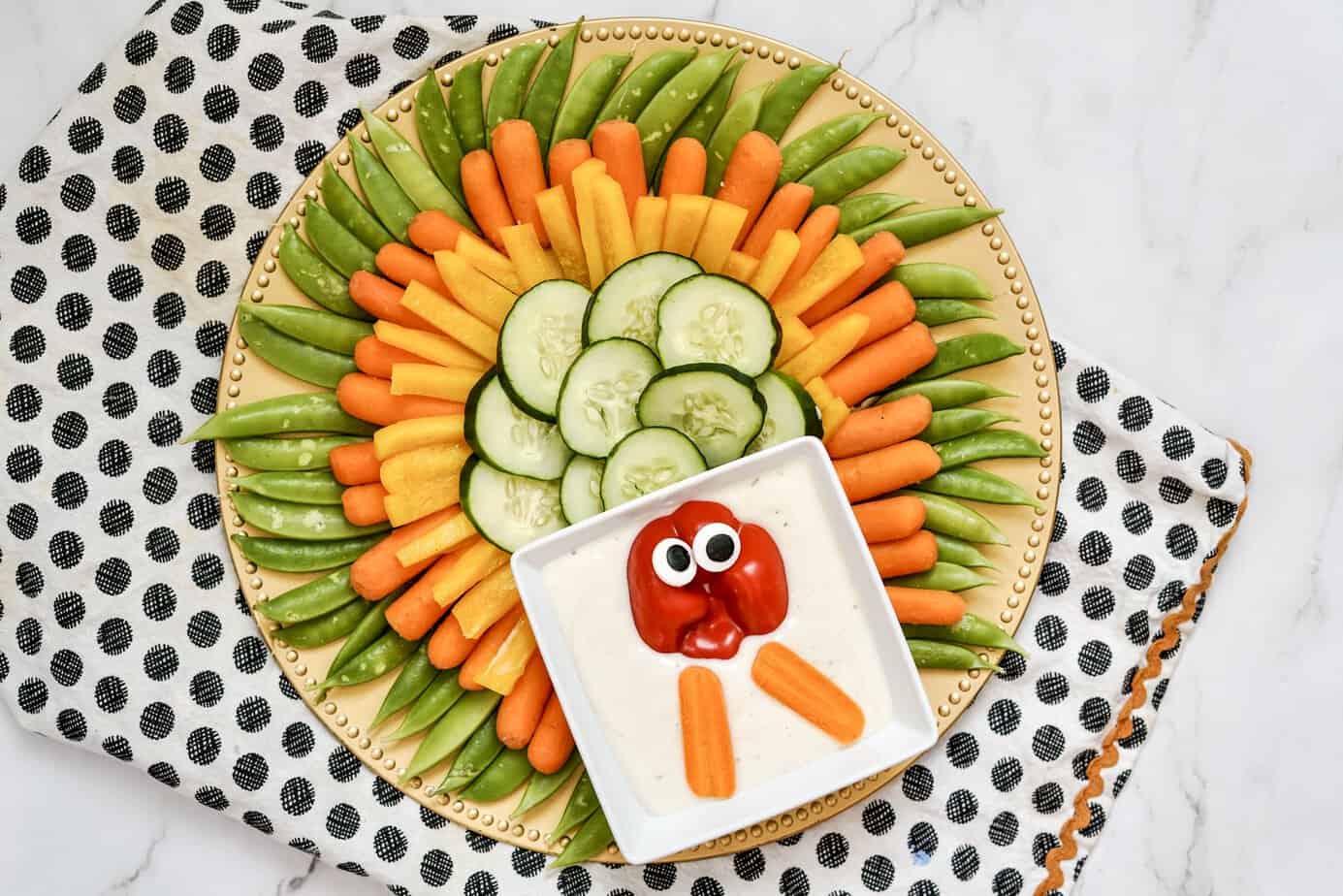A round platter with sliced cucumbers, carrots, yellow carrots, and snap peas arranged in a sunburst pattern, with a dip decorated to look like a face using a tomato, carrots, and olives.