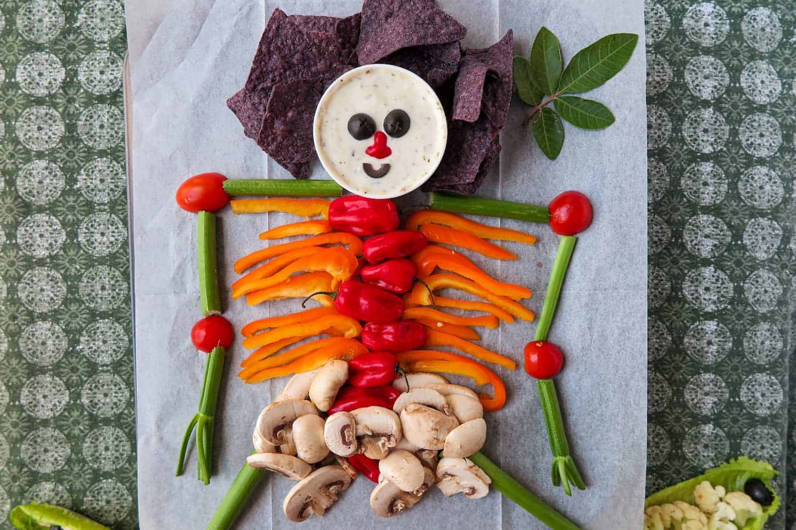A skeleton shape made from assorted vegetables and chips, with a dip cup for the head, arranged on parchment paper on a patterned green tablecloth.