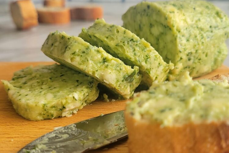A log of herb butter is sliced on a wooden board, with a knife and a piece of bread in the foreground and more bread slices in the background.