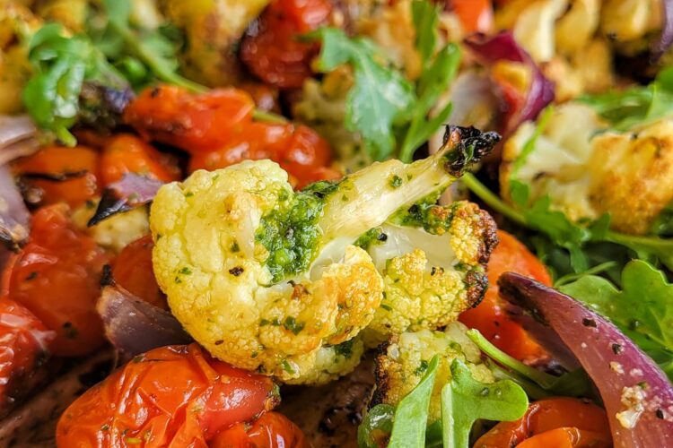 Close-up of roasted cauliflower, cherry tomatoes, red onion, and arugula, seasoned with green pesto sauce.