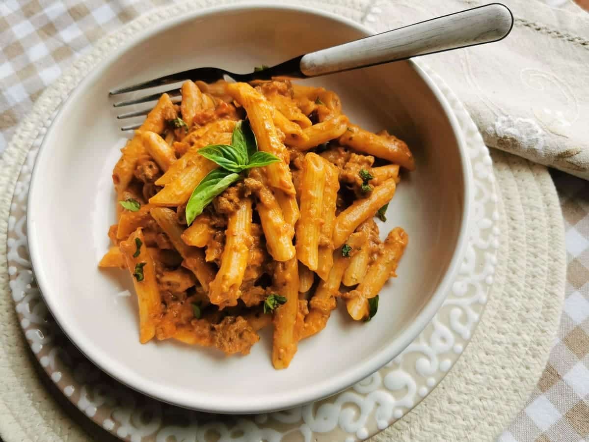 A bowl of penne pasta with meat sauce, garnished with fresh basil, served with a fork on a white plate atop a patterned placemat.
