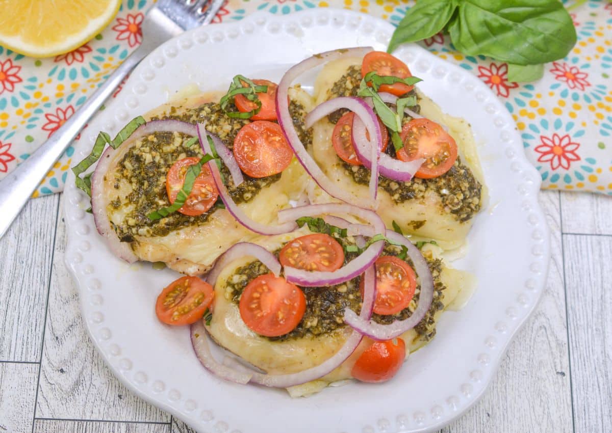 Three baked chicken breasts topped with pesto, sliced cherry tomatoes, red onion, and fresh basil on a white plate, set on a patterned napkin.