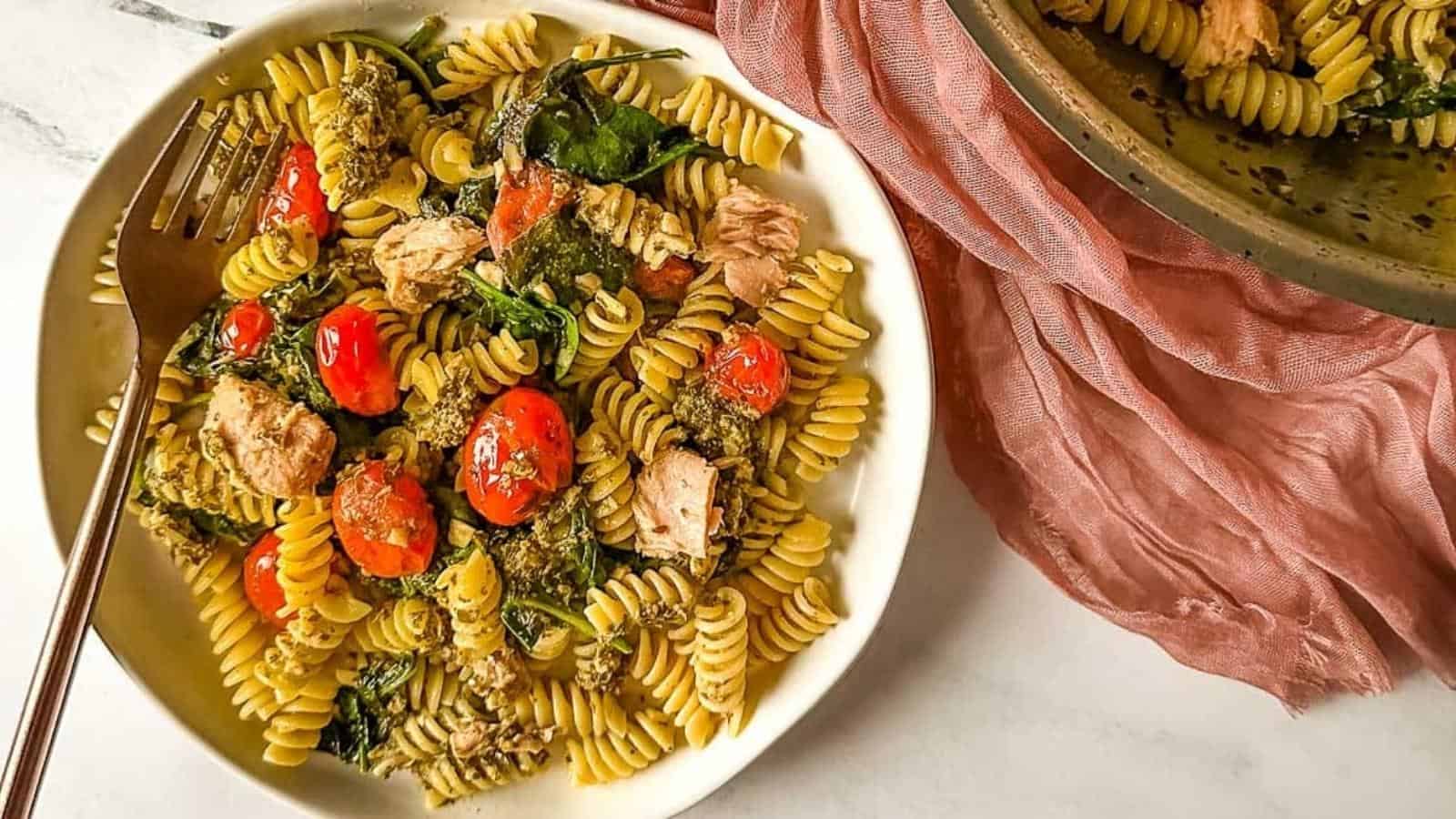 A bowl of rotini pasta mixed with cherry tomatoes, spinach, mushrooms, and chunks of tuna, with a fork on the side.