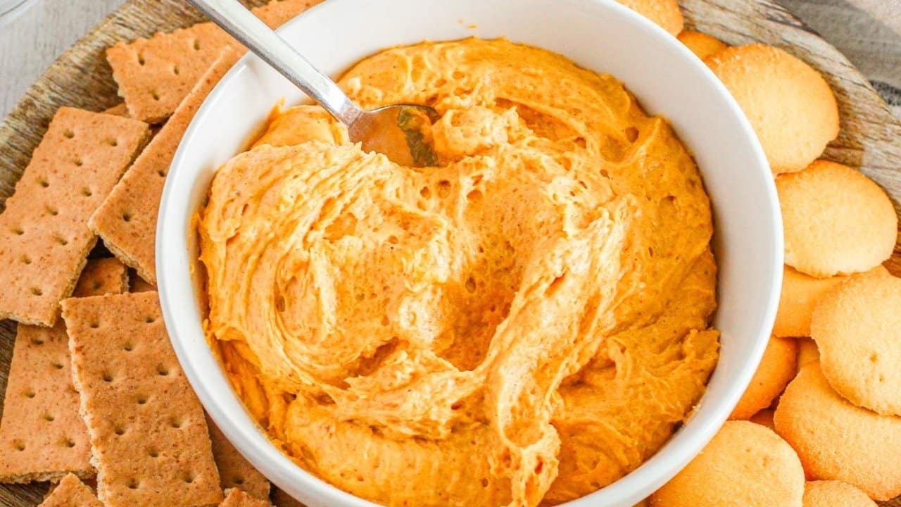 A bowl of orange dip with a spoon, surrounded by graham crackers and vanilla wafer cookies on a wooden platter.