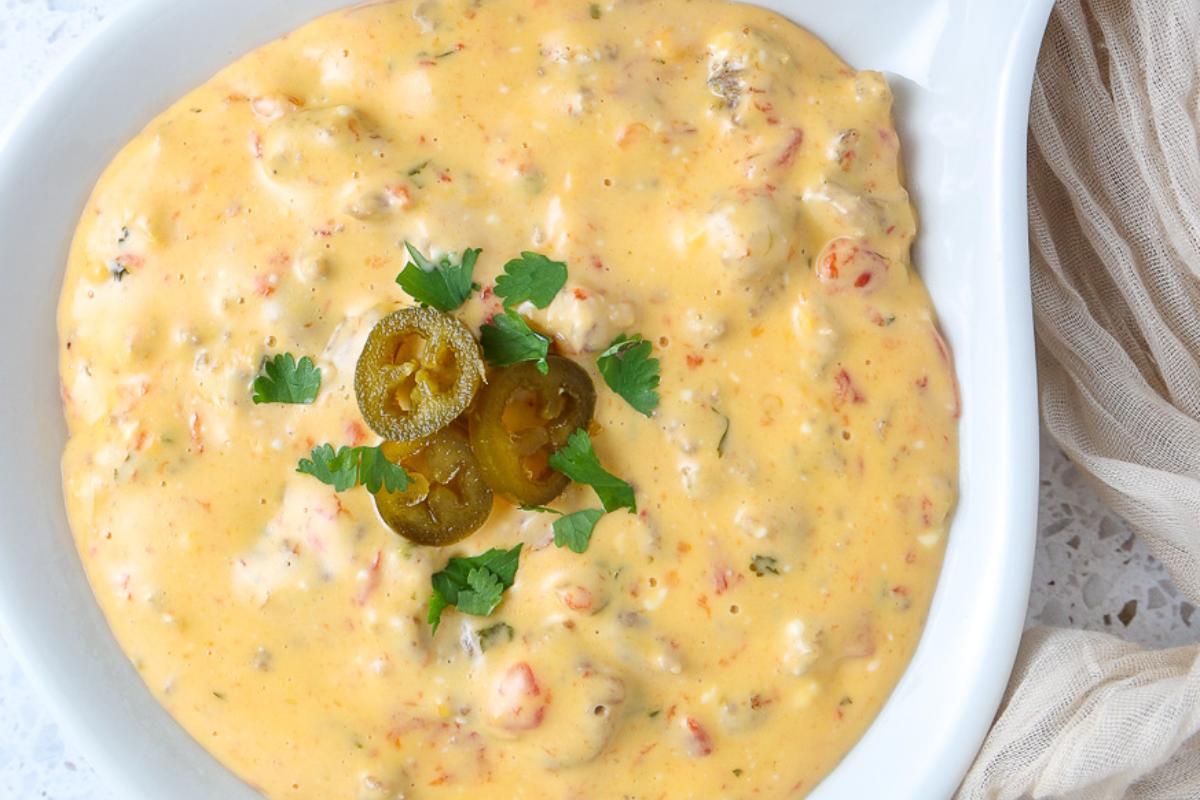 A bowl of creamy yellow queso dip with visible tomato and herb pieces, garnished with sliced jalapeรฑos and cilantro.