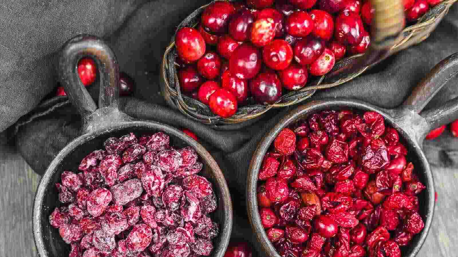 Three containers filled with fresh cranberries, dried cranberries, and sugared dried cranberries on a dark fabric background.