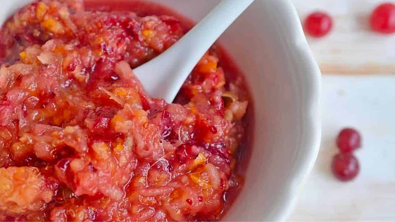 A close-up of a white bowl filled with cranberry relish, showing a white spoon and a mix of finely chopped cranberries and orange.