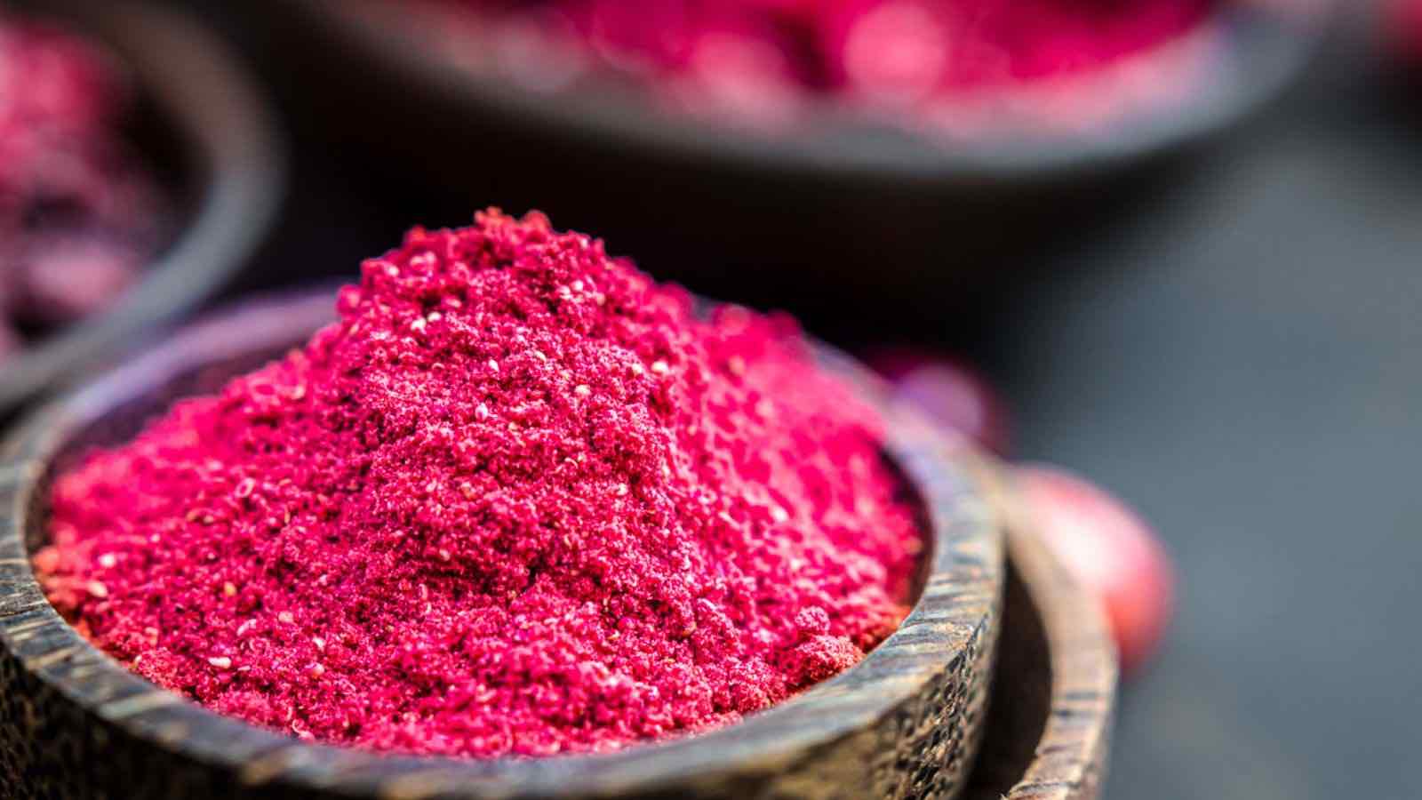 A close-up of bright red powder in a wooden bowl, with more powder visible blurred in the background.