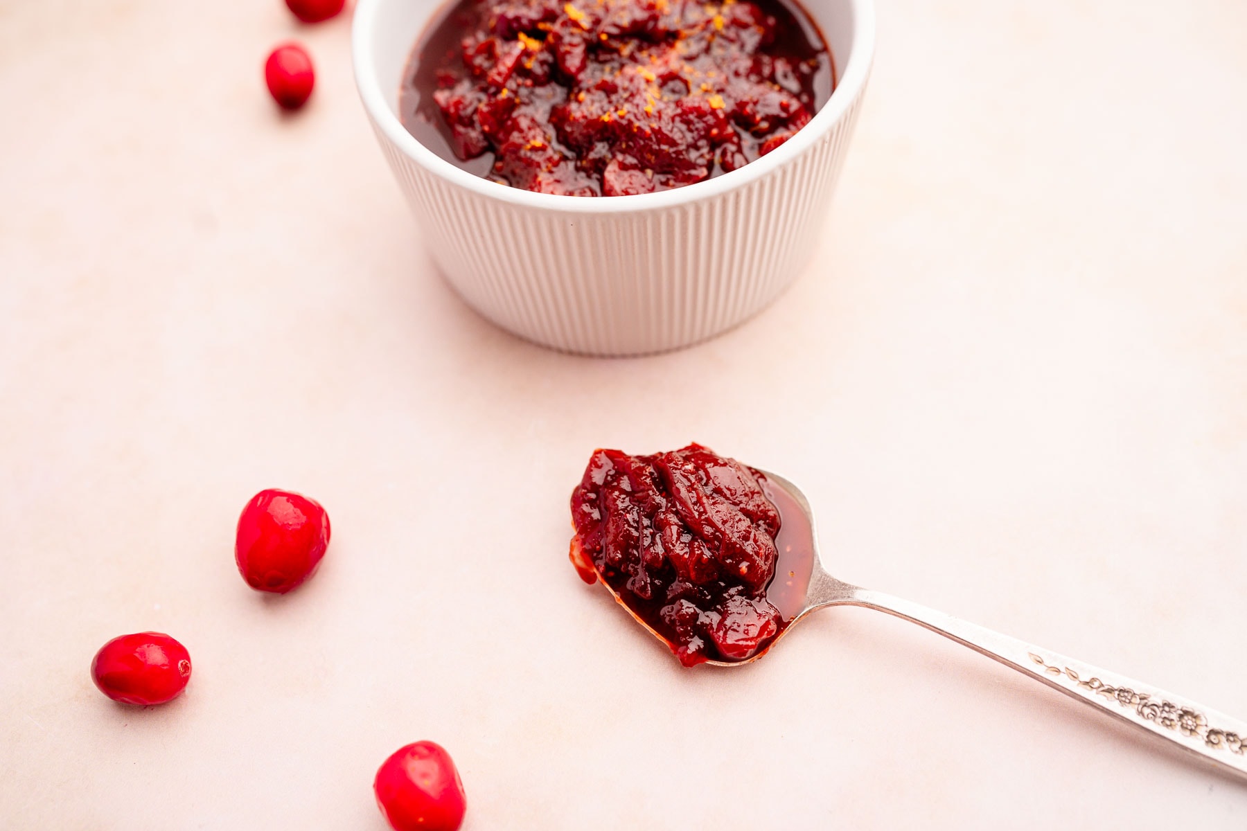 A white ramekin filled with cranberry sauce sits on a light surface, with a spoonful of sauce and a few whole cranberries nearby.