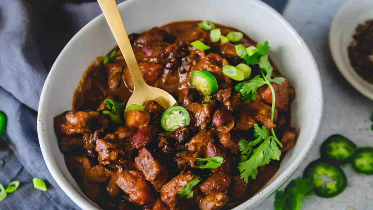 A bowl of chili with beans and meat, topped with sliced jalapeรฑos, chopped green onions, and fresh cilantro, with a gold spoon in the bowl.