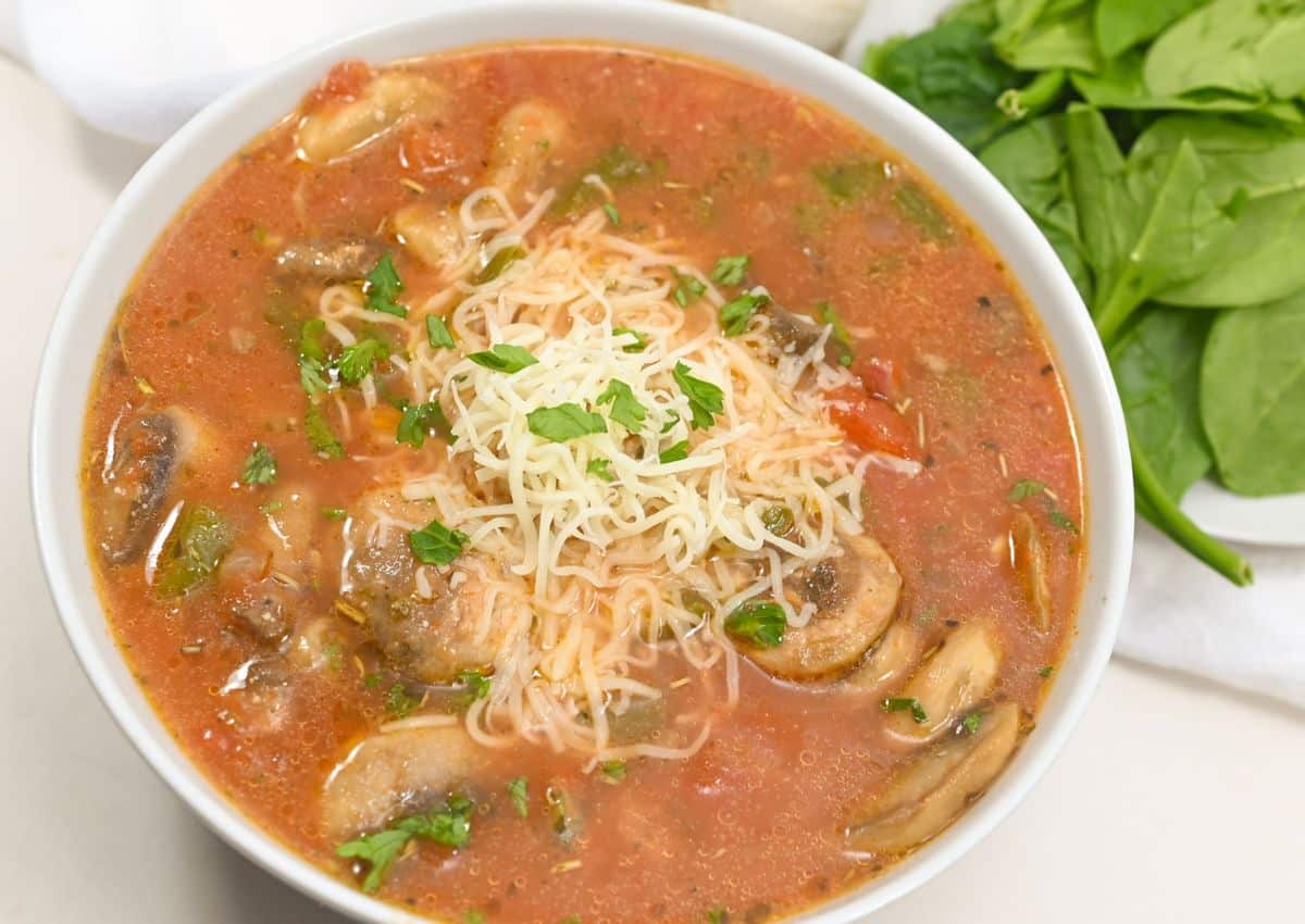 A bowl of tomato-based soup with sliced mushrooms, topped with shredded cheese and chopped herbs, next to fresh spinach leaves.