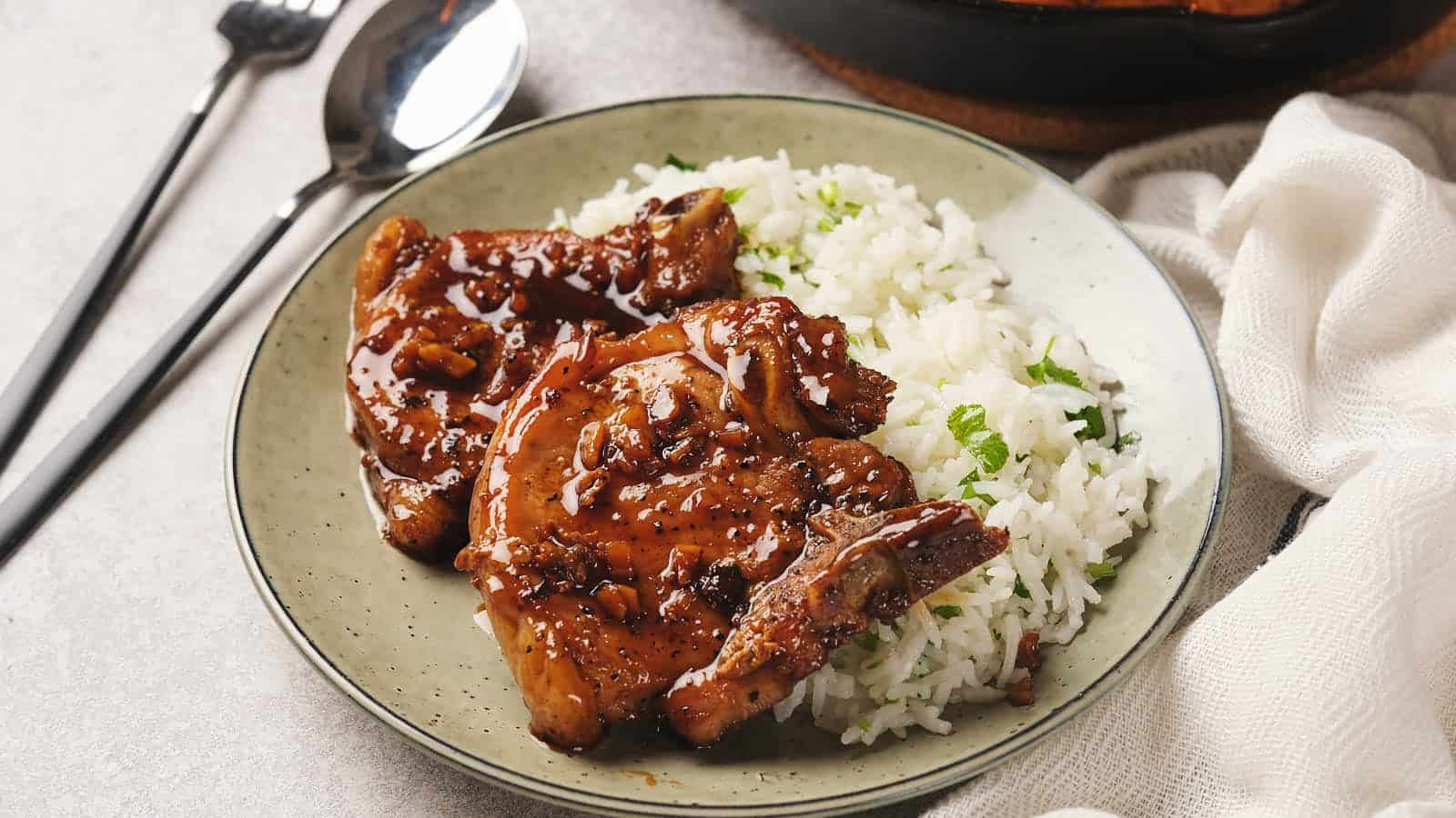 A plate of glazed pork chops served with white rice garnished with chopped herbs, next to a spoon, fork, and white cloth napkin.