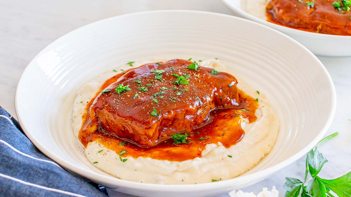 A piece of meatloaf with brown sauce sits on a bed of mashed potatoes in a white bowl, garnished with chopped parsley.