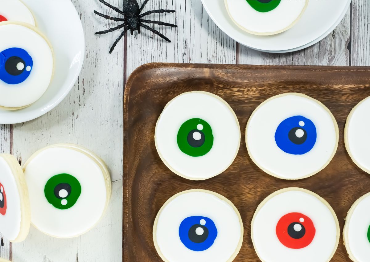 Sugar cookies decorated to look like colorful eyeballs are arranged on a wooden tray and plates, with a black plastic spider nearby on a white wooden surface.