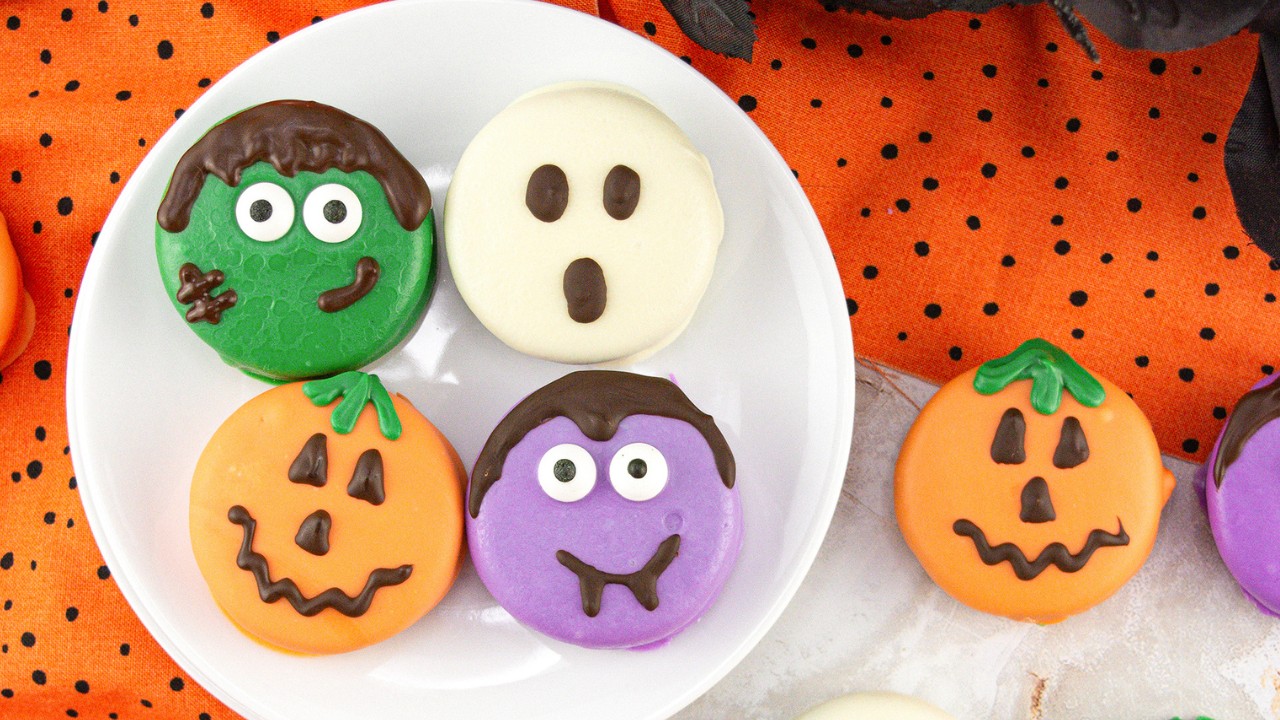 A plate of Halloween-themed cookies decorated as a green monster, a ghost, a purple monster, and two jack-o-lanterns, placed on an orange polka dot cloth.