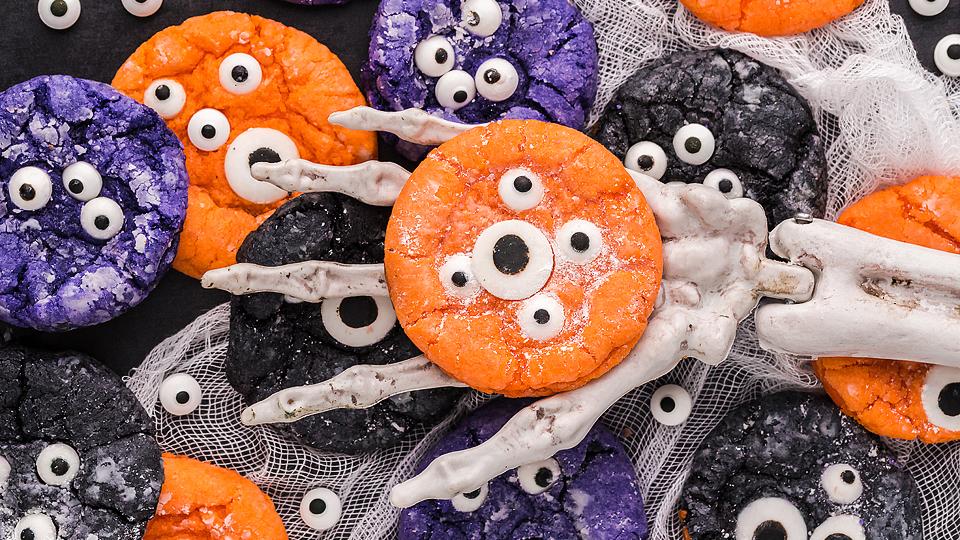 Assorted orange, purple, and black cookies decorated with candy eyes are held by a plastic skeleton hand on a white gauze background.