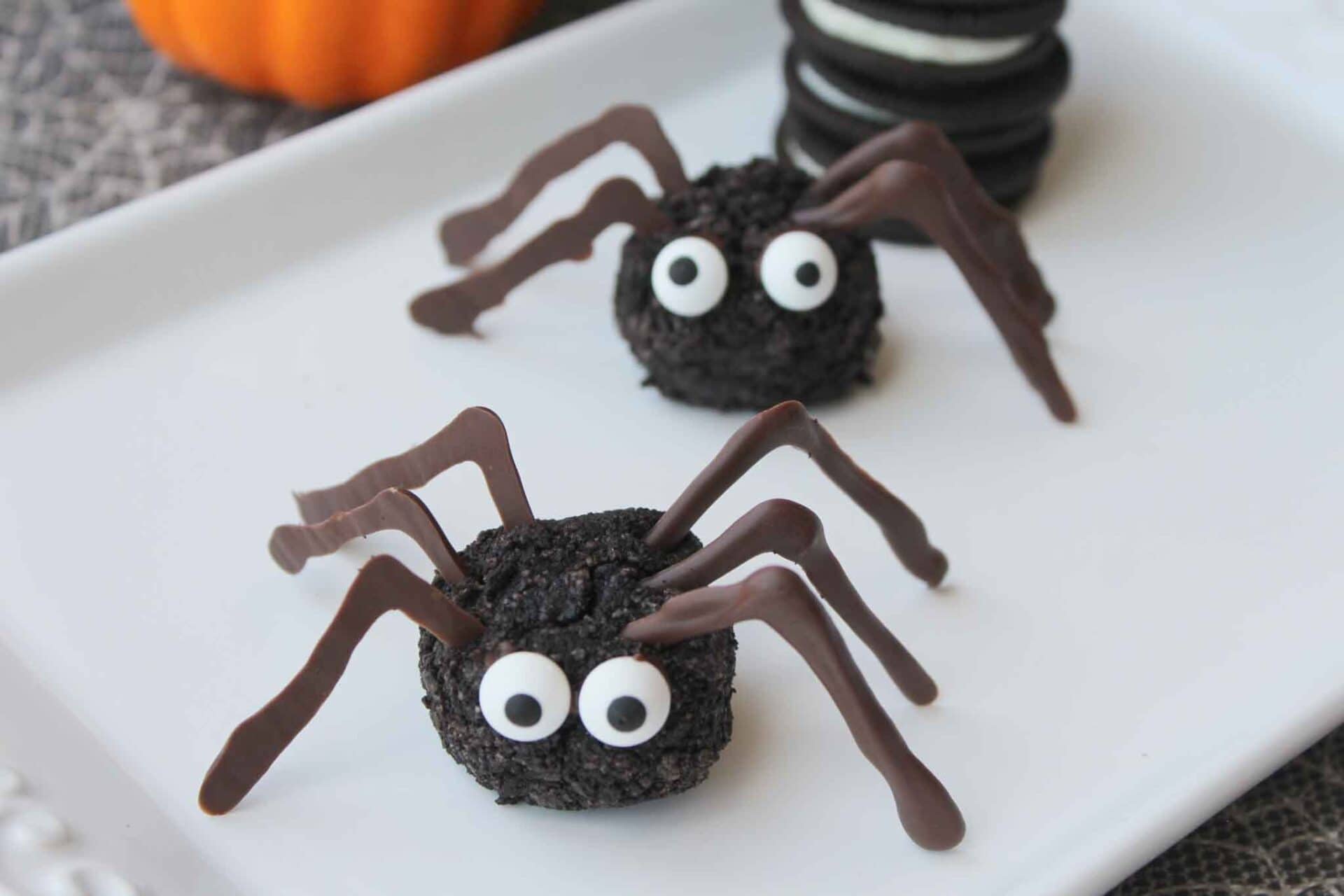 Two spider-shaped treats made from crushed cookies and candy eyes with chocolate legs are displayed on a white plate, with a stack of sandwich cookies and a small pumpkin in the background.