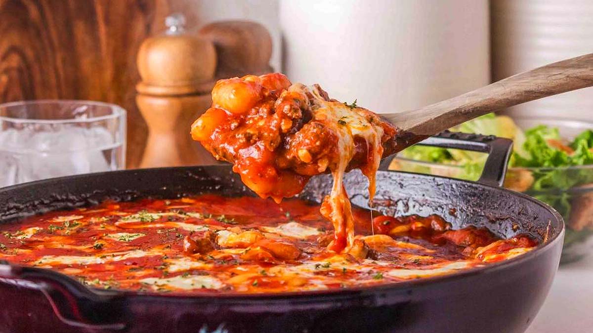 A wooden spoon lifts cheesy gnocchi in tomato sauce with ground meat from a cast iron skillet.