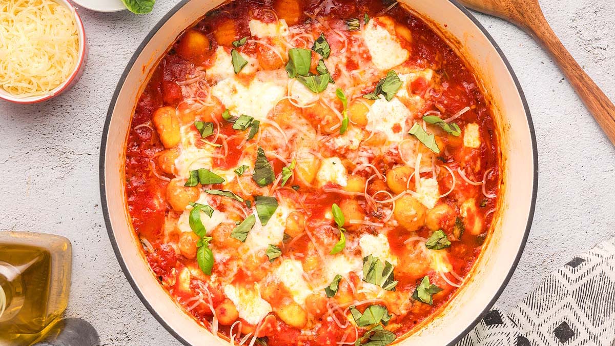 A pan of baked gnocchi in tomato sauce topped with melted cheese and fresh basil, with a bowl of grated cheese and a wooden spoon nearby.