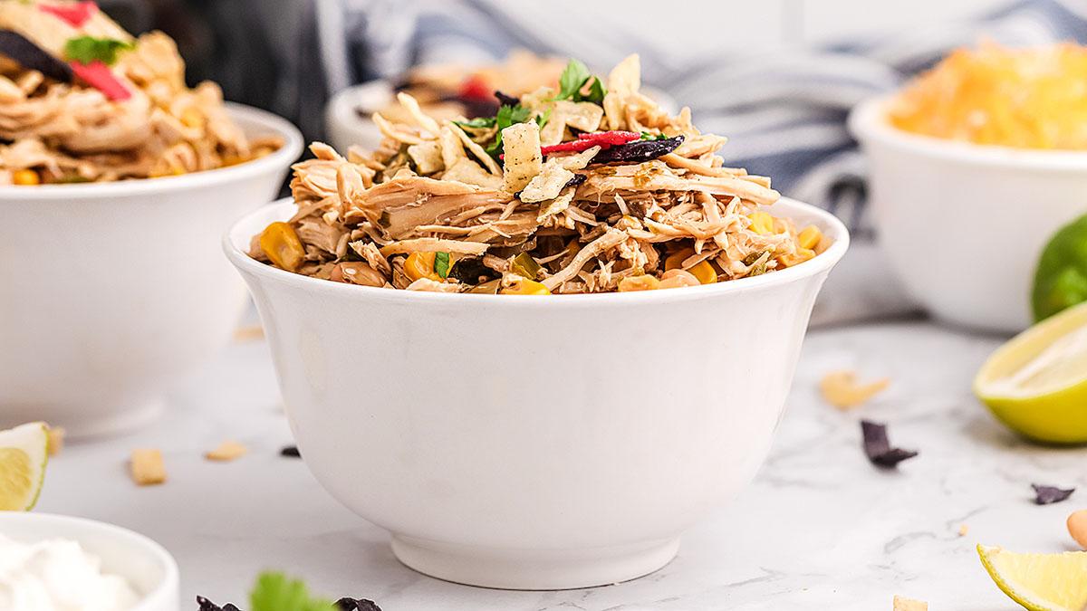 A white bowl filled with shredded chicken, vegetables, and tortilla strips on a light-colored table with other bowls and lime halves in the background.