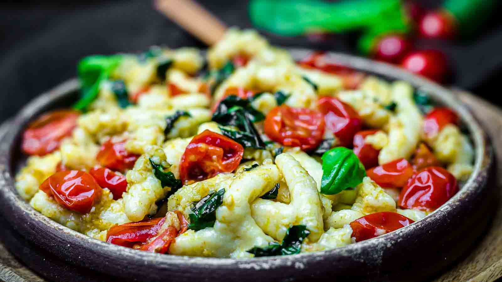 A bowl of pasta topped with cherry tomatoes, fresh basil leaves, and spinach, served in a rustic dish.