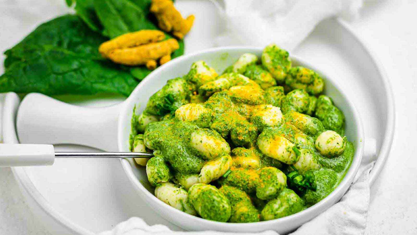 A white bowl of gnocchi covered in green sauce and sprinkled with yellow seasoning, with spinach leaves and turmeric roots on the side.